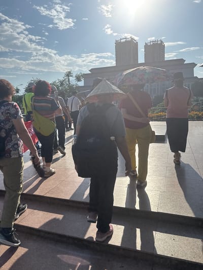 The image shows a group of people, likely tourists, in what appears to be an urban setting. Several individuals are wearing conical hats, and one person is holding a colorful umbrella. The group is standing on a paved surface that casts strong shadows, suggesting bright sunlight. Tall buildings are visible in the background, along with a blue sky dotted with clouds. The location is identified as Lao Cai Ward, Vietnam. The overall scene suggests a tour or gathering of people in a public space.