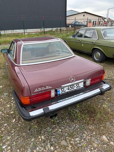 An exterior, high-angle, medium shot captures a burgundy Mercedes-Benz 450 SL parked outdoors on a gravel lot. The car faces right, its rear bumper and taillights prominently displayed. A license plate with the text "SL-450-SL" and the country code "GE" for Georgia is visible. To the right of the Mercedes is a section of a green Mercedes-Benz sedan. In the background, a dark grey building with a corrugated metal facade is to the left, and a lighter colored building with windows and an arched entryway is situated further back and to the right. Fencing separates the foreground from the background buildings. The ground is covered in loose gravel and sparse vegetation, with a few small yellow wildflowers visible. The sky is overcast.