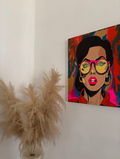 The image depicts an interior space, possibly a residential or commercial setting, likely in Rumuokwuta, Nigeria (as requested in the prompt). A painting featuring a stylized portrait of a woman wearing round sunglasses with green lenses and pink frames hangs on the wall. The woman has dark hair, tan skin, and red lipstick. The background of the painting is a mixture of colors, including red, gold, blue, and pink, with a swirling, fluid effect. Below the painting, a vase containing a cluster of tan-colored pampas grass is present. The walls are white. No people are directly visible in the scene, the only subject being the painting. The interaction is between the wall, the painting and the vase with the pampas grass.