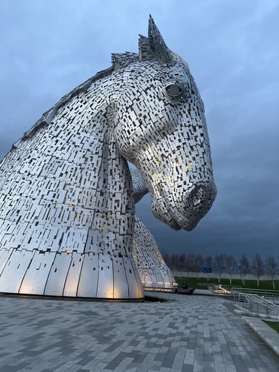 The image displays one of 'The Kelpies' monumental horse head sculptures, located in Helix Park, Falkirk, United Kingdom, under an overcast sky. The sculpture is constructed from numerous interlocking rectangular and irregular brushed metal panels, presenting a silver-grey appearance. Warm yellow light emanates from internal sources through gaps in the plating and from ground-level spotlights illuminating its base. The horse's head is angled downwards and to the right, featuring a distinct circular eye detail. The foreground consists of a grey paved pathway composed of rectangular pavers. In the midground, a grassy bank rises, populated by a line of leafless deciduous trees and bordered by a metal railing. No individuals are visible, and no discernible actions or interactions are occurring within the scene.