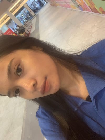 An indoor, horizontally oriented selfie features a young woman with long, dark hair, wearing a blue top, positioned in the foreground. Her head and upper torso dominate the frame, angled slightly upwards. She has a neutral expression, with subtle makeup on her eyes and lips, and a small blemish visible on her chin. A black strap, possibly from an undergarment, is visible beneath her blue top on the right side of the image.

The background is blurred, indicating an indoor public setting, likely a shopping mall in Samarinda, Indonesia, given the context. The floor appears to be light-colored tile. To the far left, a colorful, abstract graphic or poster is partially visible. In the lower left quadrant of the background, reflections and blurry elements suggest distant displays or storefronts with artificial lighting. Fragments of text are barely discernible, with "LE" being somewhat visible on a red background in the distance. The overall lighting suggests an artificial indoor environment. The woman is the sole clear subject, engaged in the act of taking a self-portrait.