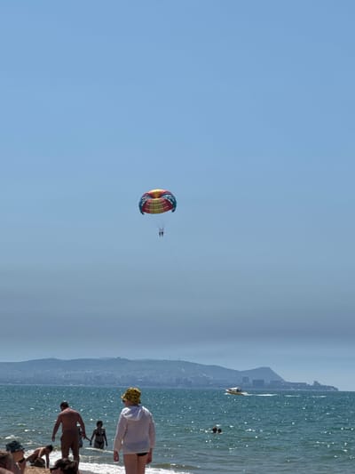 In Vityazevo, Russia, on a bright, clear day, two individuals are parasailing high above the Black Sea. Their colorful parachute, a mix of red, yellow, and blue, is a vibrant contrast against the pale blue sky. Below them, the sea is a deep blue, with small whitecaps indicating a gentle breeze. A narrow strip of sand is visible along the shoreline, with several people wading in the shallow water. In the distance, a city sprawls along the coast, nestled at the foot of rolling hills that fade into a hazy horizon. A small motorboat cruises across the water, leaving a white wake in its path. The scene captures a typical summer day at a popular beach resort.