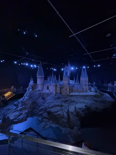 This media file captures a nighttime scene of a detailed model of a castle, likely Hogwarts from the Harry Potter series, set against a dark, possibly theatrical, background. The model is illuminated with blue and yellow lights, highlighting the intricate architecture with its multiple spires, towers, and windows. It sits atop a rocky, uneven terrain that appears to be part of the exhibit.
In the foreground, thin wires or cables stretch across the frame, suggesting the model might be suspended or part of a larger display. Scattered across the dark background are small, bright blue lights, resembling stars or a starry sky. On the right side of the image, a metal railing or barrier is visible, implying that viewers are observing the model from a designated area.
The overall ambiance is mysterious and enchanting, enhanced by the low lighting and the majestic, albeit miniature, castle. The blue lighting creates a magical, almost ethereal atmosphere. There is no visible text in the image. Given the context of the United Kingdom, the subject matter strongly points to a Harry Potter-themed attraction, possibly within a film studio tour or theme park. The lack of people and the nighttime setting contribute to the focus on the elaborate model itself.