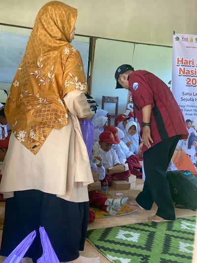 In Handil Baru, Indonesia, an indoor scene unfolds with a group of young children dressed in school uniforms, predominantly red and white, seated on a green and white patterned rug. Some children are wearing red hats. A man in a dark red shirt and black pants, wearing a black baseball cap, is interacting with the children. He appears to be handing out or distributing items. To the left of the frame, a woman in a gold hijab and a light-colored tunic stands with her back to the camera, partially obscuring a purple plastic bag. The room has a rustic feel with visible wooden beams and paneling on the walls. A banner with text in Indonesian, including "Hari" and "Nasional," is visible in the background, suggesting an event or celebration. The floor is tiled, and a dark duffel bag is placed to the right. The overall atmosphere suggests a charitable event or educational outreach program for children.