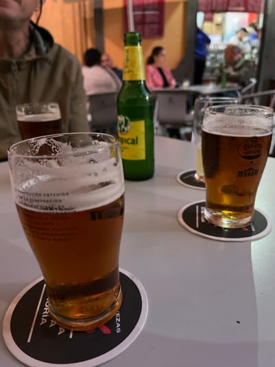 The image shows a table in a bar or restaurant in Arucas, Spain, located at the coordinates 28.118 latitude, -15.524 longitude. The table is covered with a white tablecloth. On the table, there are two glasses of beer, one with a white head, both sitting on black circular coasters. The beer in the foreground is a light amber color and is in a clear glass. There is a green glass bottle with a label that reads "Tropical," behind the glass in the foreground. The bottle is mostly empty, with only a small amount of liquid remaining. The background of the image is blurred, but it appears to be a bar or restaurant with people seated at tables. The scene appears to be typical of an outdoor cafe in the city of Arucas.
