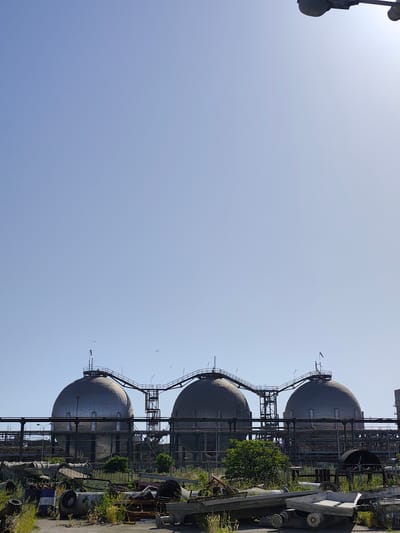 The image shows a clear blue sky above a row of three large, spherical industrial storage tanks. These tanks are connected by a network of elevated walkways and pipes. In the foreground, there is a cluttered area with various pieces of scrap metal, barrels, and machinery, overgrown with grass and weeds. Some of the barrels appear to have labels, but they are not legible. The overall scene suggests an industrial setting, possibly a storage facility or a decommissioned industrial site. The lighting indicates it is daytime, and the clear sky suggests fair weather. There are no people visible in the image. The presence of industrial infrastructure and scrap materials are the main subjects.