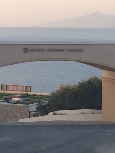 An arched, light-colored structure spans across the upper-middle section of the image, prominently displaying the text "MITSIS SUMMER PALACE" in dark letters, preceded by an emblem resembling a sunburst or floral motif. The scene, identified as being in Pyli, Greece, is viewed through this archway. In the immediate foreground, a dark gray paved road surface is visible. Beyond this, descending terrain features a light-colored, curved retaining wall on the left, and a partial view of a beige building on the right. Below the arch, a diverse landscape includes various green shrubs, low-lying vegetation, and sections of stone-faced and white plaster walls. Near the water's edge on the left, a linear, multi-paneled wooden structure, possibly a barrier or deck, is present. The midground is dominated by a calm body of water, likely the Aegean Sea, extending horizontally across the frame. In the distant background, a hazy, light-colored landmass, appearing as mountains or hills, is faintly visible against a pale sky. No individuals, direct actions, or interactions are depicted. The lighting is soft and diffuse, suggesting either early morning or late afternoon conditions.