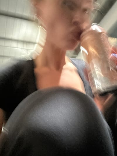This heavily motion-blurred, low-angle image captures a person in motion, possibly indoors. The main subject, a person with fair skin, is visible on the left side, with their head and face highly blurred. They appear to be holding a clear glass or bottle, which prominently reflects bright light, possibly bringing it towards their mouth.
The person is dressed in dark clothing, likely black or dark grey, visible on the right side of the frame, possibly a shoulder or upper arm. A section of lighter grey or blue clothing can also be seen further towards the center, suggesting torso wear.
The background is indistinct due to the motion blur but contains blurry vertical or horizontal lines, indicative of an interior setting such as a corridor, a room with blinds, or possibly the interior of a moving vehicle. The extreme blur implies significant movement or camera shake during capture. No specific emotions are discernible due to the blur. The time of day and weather cannot be determined from the image. There is no visible text. The location context of Tonbridge and Malling, United Kingdom, is not visually identifiable within the image.