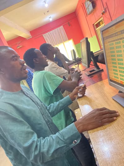 This image depicts an interior scene within a computer facility or internet cafe in Chikaji, Nigeria. Four adult males are visible, positioned along a continuous wooden counter. The room features reddish-orange walls, a light-colored ceiling with two illuminated bare light bulbs, and white-curtained windows in the background. The closest male, wearing a light teal patterned long-sleeved garment, holds a small white paper slip in his right hand while resting both hands on the wooden counter, his gaze directed forward towards a computer monitor. This monitor displays a grid of green squares with a "00:41" timer. Behind him, a male in a green t-shirt has his hands clasped on the counter, also looking forward. Further down the counter, a third male in a light-colored shirt is visible, with one hand touching his chin as he looks towards a screen. A fourth individual is partially visible. Multiple black-framed computer monitors are present on the counter.