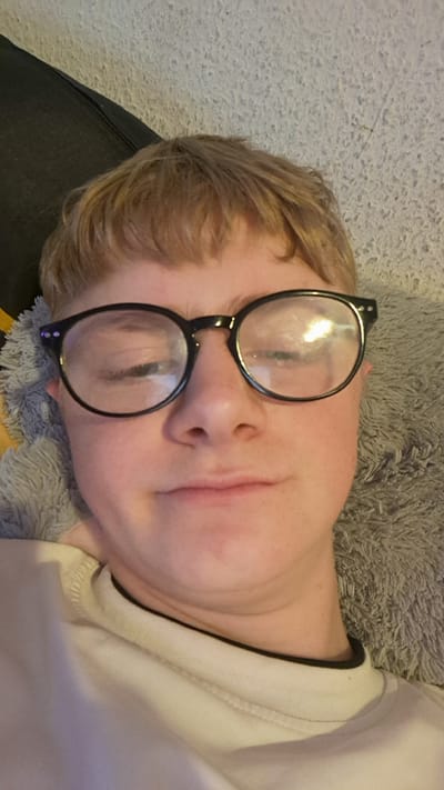 The image is a close-up, selfie-style portrait of a young person wearing black-rimmed glasses. Their hair is light brown and styled with bangs. The background appears to be a textured surface, possibly a carpet or blanket, in a light gray color. A dark surface, possibly furniture, is visible in the upper left corner. The lighting in the image is dim, with reflections visible on the glasses lenses. The person's expression is neutral, with one eye slightly closed. The image does not contain any information that would allow for the determination of its location in the city of Parchim, Germany.
