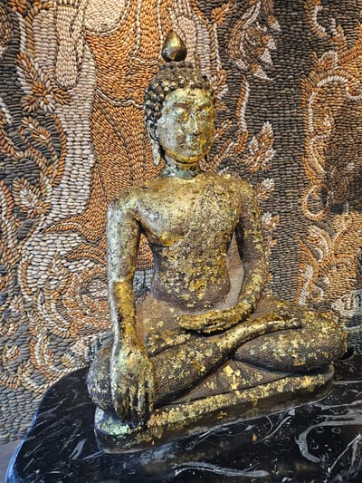 A seated Buddha statue, gilded with gold leaf, is positioned in front of a wall mosaic made of small, smooth pebbles. The mosaic depicts an abstract pattern in shades of brown, beige, and gray, resembling flowing organic forms. The Buddha statue itself is dark gray underneath the gold leaf, which is unevenly applied, with some areas of gold being more prominent than others. The statue has a serene expression and is seated in a lotus position. It rests on a dark, polished surface, possibly marble, with white veining. The background mosaic is highly detailed and covers the entire wall behind the statue. The lighting is soft, casting subtle shadows. This scene is located at the Sanctuary of Truth in Pattaya, Thailand, which is near Na Chom Thian.