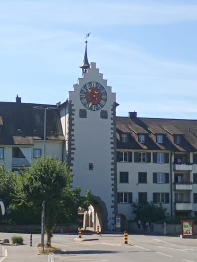 A high-angle, full shot captures a white clock tower with a pointed roof and a weather vane, set against a clear blue sky. The tower is adorned with a large, colorful clock face showing the time as approximately 3:50. Below the clock, a dark arched gateway opens into a shadowed area, where several people stand. The clock tower is flanked by buildings with dark roofs and multiple windows. To the left of the tower, a section of a lighter-colored building with a dark, sloped roof is visible, featuring a balcony with a white railing. To the right, a multi-story residential building with several balconies is present. In the foreground, a street intersection is shown, with a pedestrian crossing marked by white lines. Yellow and black bollards line the intersection, and a street lamp stands near the center of the frame. Trees and shrubs are scattered throughout the scene. The overall scene depicts a street in the historic town of Stein am Rhein, Switzerland.