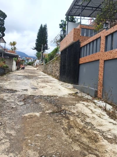 A narrow, dilapidated street ascends slightly, flanked by walls and buildings. The road surface is severely damaged with potholes and uneven surfaces. A modern-style gate with dark vertical slats is on the right, while a stone wall runs along the left. In the distance, houses and trees are visible under a cloudy sky. A parked motorbike sits further up the street. The overall scene is one of neglect and disrepair.