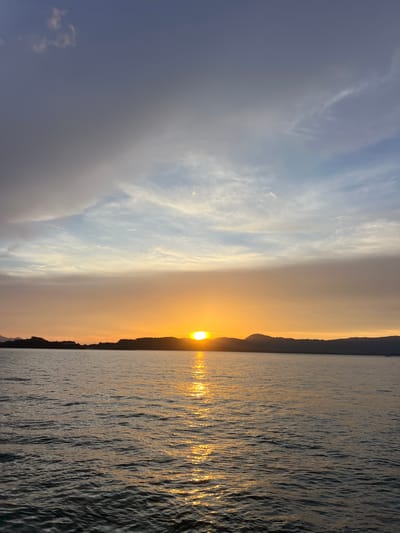 The image depicts a serene sunset over a body of water, likely a bay or lake, with a silhouetted coastline in the distance. The sun is partially visible, casting a golden-orange glow that reflects vibrantly on the rippling water's surface. Above the horizon, the sky transitions from warm sunset hues to cooler blues, with wispy clouds scattered across the expanse. The overall impression is one of tranquility and natural beauty. The location is identified as Pakhlok, Thailand. There are no people or specific objects or actions to describe.