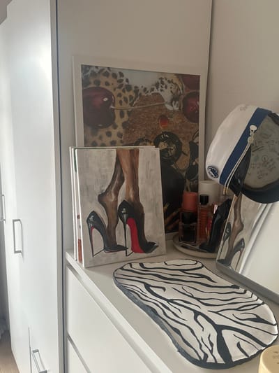 The image shows a dresser with several decorative items on top. There is a framed artwork of cherries and a leopard print, a painting of a woman's legs in high heels, several perfume bottles on a tray, and a decorative tray with a zebra print pattern. A white hat with a blue and gold trim is also visible. The dresser is white and has silver handles. The background shows a white wall. The scene is indoors, likely a bedroom or dressing area. The location is identified as Varde, Denmark. There are no people present in the image.