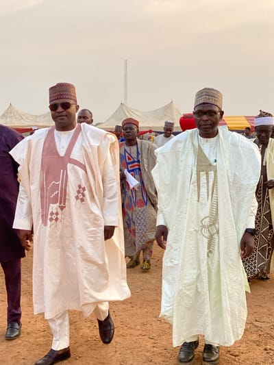 The image depicts a group of men dressed in traditional attire, likely attending an outdoor event. The men wear flowing robes, some with intricate embroidery. Headwear varies, with some wearing caps or hats. The ground appears to be sandy, and tents are visible in the background. The sky is overcast, suggesting a hazy or cloudy day. The men are standing in rows, possibly posing for a photograph.