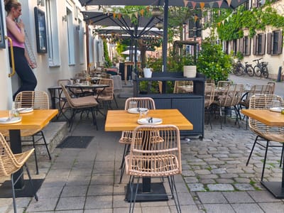The image depicts an outdoor seating area of a restaurant or café, likely located on a cobblestone street in Erlangen, Germany. In the foreground, several tables with wicker chairs are arranged. One table, made of light-colored wood, is set with white plates, cutlery, and glasses. A woman, wearing a pink tank top and black pants, stands by a building wall on the left side of the frame, holding a long glass bottle. The background shows more tables and chairs under umbrellas, as well as a row of buildings with shuttered windows, some of which are covered in ivy. Two bicycles are parked against one of the buildings. The overall scene suggests a pleasant, relaxed atmosphere typical of a European town center.
