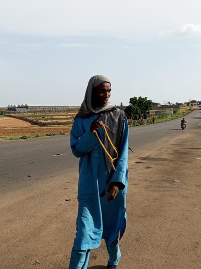 A man in a blue robe and head covering walks on the side of a road in Gadao, Nigeria. He is holding a string of yellow beads in his right hand. The road is paved and appears to be a main thoroughfare, with a few vehicles and pedestrians visible in the distance. The landscape around the road is dry and dusty, with some sparse vegetation and agricultural fields. In the background, there are several large, modern buildings that appear to be industrial or agricultural structures. The sky is clear and blue with a few scattered clouds. The overall scene suggests a rural or semi-rural setting in Nigeria.
