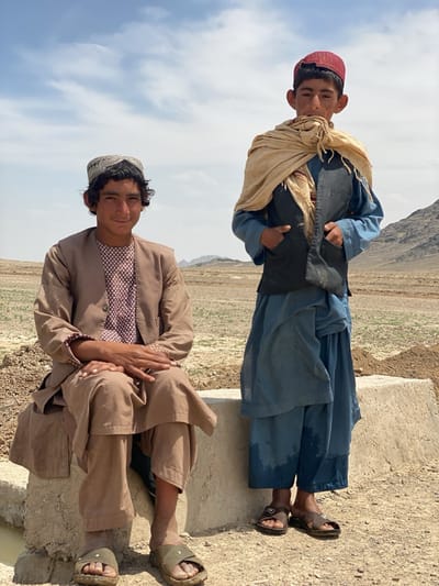 Two young males are depicted in a full-length, outdoor portrait against an arid landscape in Ashoqah, Afghanistan. The bright, clear daylight suggests it is midday or early afternoon, with a few clouds visible in the pale blue sky.
On the left, a young male sits comfortably on a light-colored concrete block. He wears a traditional brown garment consisting of a long-sleeved coat over a patterned, perhaps maroon and white, collared shirt. A light-colored, embroidered cap covers his dark, curly hair. He looks directly at the camera with a gentle, friendly smile, his hands clasped casually on his lap. He wears simple, open-toed sandals.
Standing to the right, beside the concrete block, is another young male. He is dressed in a loose-fitting, light blue traditional outfit, possibly a shalwar kameez, with a dark, sleeveless vest over it. A long, light beige scarf is wrapped around his neck and shoulders. A red cap sits on his head. He has a more neutral, perhaps slightly pensive expression, looking towards the camera, with his hands tucked into the pockets of his vest. He also wears dark, open-toed sandals, and the lower part of his trousers appears damp or stained.
The setting is a dry, rural area with sparse green vegetation in the foreground and distant, rugged mountains under a hazy sky. The ground is sandy and dusty. The concrete structure they are near could be part of an irrigation system or a rudimentary bench. There is no visible text in the image.