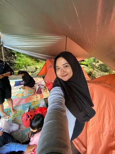 Inside a tent with an orange exterior, a woman in a black hijab takes a selfie. Below, a colorful cartoon-themed mat is spread out, upon which several people are relaxing. Some are seated, while others are lying down. Red plastic bags and assorted items are scattered on the mat. A sliver of greenery is visible just beyond the tent's entrance.