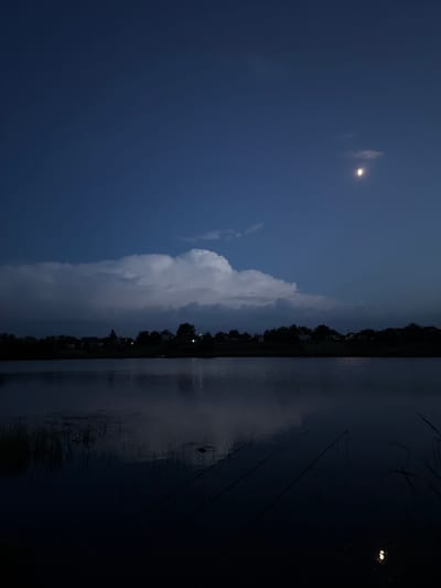 The image depicts a nighttime scene at a body of water, likely a lake or river, near a settlement in Hrebinky, Ukraine. The sky is a deep blue, with a sliver of the moon visible and a large cloud formation dominating the middle ground. Silhouetted against the sky is a line of houses and trees along the far bank of the water. The water itself is dark and reflective, with some reeds or grasses growing along the near edge. Several fishing rods are visible, set up along the bank, suggesting an activity in progress. The overall atmosphere is tranquil and somewhat eerie due to the low light conditions.