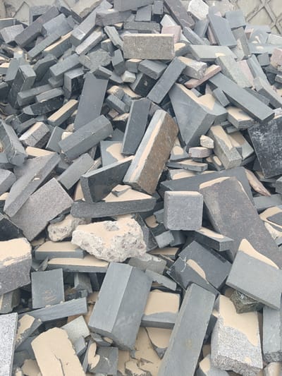 The image depicts a large pile of irregularly shaped and sized stone or concrete debris, possibly from a construction or demolition site. The color palette consists predominantly of gray, black, and beige hues. The debris includes rectangular, block-like pieces, with a mix of smooth and rough textures. Some pieces show internal fracturing or degradation. A light-colored, fine particulate matter, possibly sand or dust, covers much of the surface of the debris. The background shows a nondescript wall in a similar gray tone. The location, Яльгелево, Russia, cannot be determined from the visual information provided.