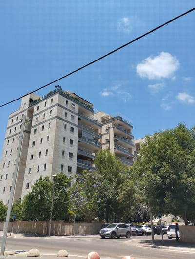An eye-level, slightly low-angle, full shot captures a tall, modern apartment building with a beige stone facade and dark gray accent stripes on each floor. The building features numerous rectangular windows and balconies with glass railings. A shorter, attached building is visible to the right, also with balconies. In the foreground, a two-lane asphalt road is depicted, with a concrete barrier separating it from a sidewalk. Several round concrete bollards are placed along the sidewalk. To the left of the road, a grey lamppost stands. Several trees with green foliage and a few trees with purple blossoms are situated between the buildings and the road. A black wire runs diagonally across the top of the frame against a blue sky with scattered white clouds. Several cars are parked in a lot behind the trees and building. The scene appears to be in the city of Pardes Hana-Karkur, Israel, based on the context provided.