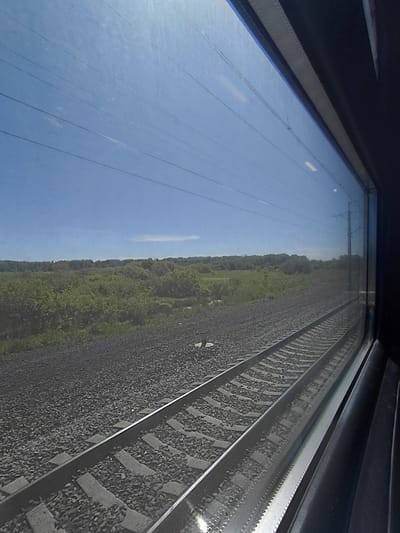 The image is taken from inside a train, looking out of the window. The scene outside shows a clear blue sky with power lines crisscrossing it. Below the sky is a vast expanse of green foliage, appearing to be fields or a wooded area, with trees and bushes. In the foreground, a set of train tracks runs parallel to the window, composed of gravel ballast and wooden ties. The tracks recede into the distance, suggesting the train is in motion. The right side of the image shows the interior frame of the train window, a dark, possibly padded, surface. The overall impression is a sunny day with a view of the countryside from a moving train. The location is identified as Kabinetnoye, Russia.