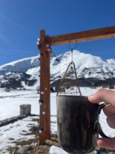 The scene depicts an outdoor winter environment in Virak, Montenegro, under a clear blue sky. In the immediate foreground, a person's right hand grasps a dark, glossy ceramic mug, which occupies the lower right portion of the frame. The mug appears to have vertical streaks, possibly from condensation or liquid. In the middle ground, a rustic wooden structure, likely a pergola or swing frame, is visible. It consists of a vertical post on the left and a horizontal beam extending across the top. A string of clear-bulbed lights is wrapped around the left vertical post. Suspended from the horizontal beam is a woven, teardrop-shaped hanging chair or swing. The ground is covered in snow, with areas of exposed dark earth or gravel. A low, stacked stone structure is partially visible further back on the left. In the background, expansive snow-covered mountains rise under a bright, cloudless sky.