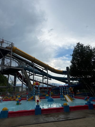 The image depicts a water park in Renens, Switzerland, under a cloudy sky. A large, multi-level water slide structure dominates the scene, with yellow, blue, and red slides descending into a shallow pool. Several children's attractions, including small slides and water features shaped like animals, are visible in the pool. A person stands at the top of one of the slides, appearing ready to descend. Trees and mountains are visible in the background. The overall impression is of a family-friendly recreational area.