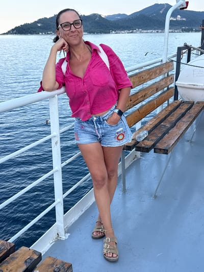 A woman wearing glasses, a pink button-up shirt, and denim shorts is standing on the deck of a ferry. She is leaning against the railing, with her left arm resting on it and her right hand in her pocket. The ferry is sailing on the water with a coastline and mountains in the background, which could be the coast of Thassos, Greece. The deck is painted a light blue, and there are wooden benches on the right side of the image. The water is a deep blue, and the sky is overcast.