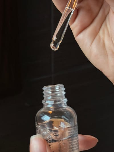 A clear, glass dropper partially filled with liquid is positioned above an open, empty glass bottle held by a hand. The bottle is lying on its side. Both items are against a stark black background. The dropper's liquid is clear. The bottle's label is partially visible, but the writing is unclear. The scene has a sterile, clinical aesthetic.