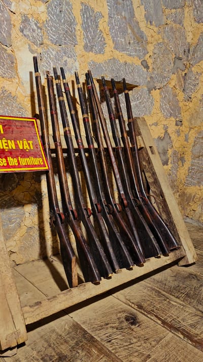 The image shows a collection of antique rifles displayed in a wooden rack against a textured wall. A sign in yellow and red lettering is partially visible on the left. The rack is situated on a wooden floor. This scene is located in the city of Sà Phìn Commune, Vietnam.