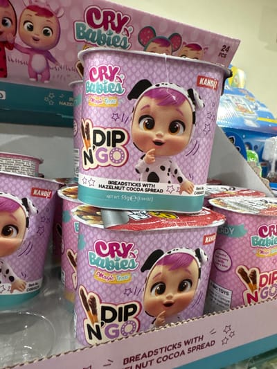 The media file shows a close-up, overhead view of several boxes of "Cry Babies Dip N'Go" snack items. The boxes are predominantly purple and feature cartoonish images of "Cry Babies" characters, which appear to be baby dolls. Each box contains breadsticks and a hazelnut cocoa spread. Visible text on the boxes includes brand names, product descriptions like "HAZELNUT COCOA SPREAD," "Magic Tears," and "DIP N'GO," as well as production details such as "Made in Türkiye" and item numbers. The items are arranged in a retail setting, likely a store shelf, with other products visible in the background, including brightly colored toy packaging. The lighting suggests an indoor environment. The overall impression is one of merchandise display for children's snacks.