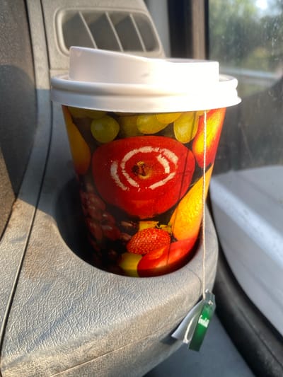 A close-up, eye-level shot shows a disposable coffee cup with a white lid, patterned with various fruits, sitting in a gray textured cup holder. A tea bag tag labeled "Greenfield" hangs over the edge of the cup. The background is blurred, showing hints of outdoor scenery, suggesting the cup is in a vehicle. The lighting suggests it is daytime. The location is identified as Changilari, Georgia, though no specific urban elements are visible.