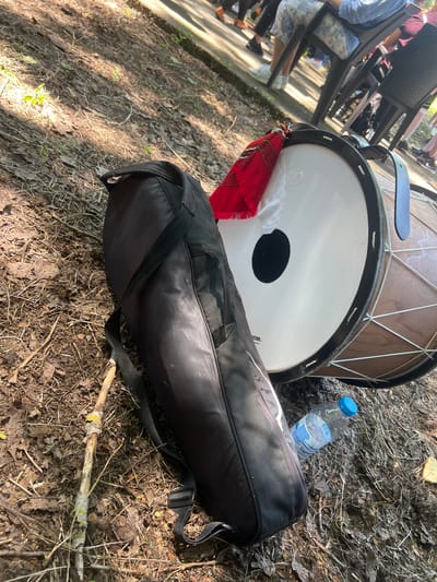 The scene depicts an outdoor gathering in Bolyarovo, Bulgaria. In the immediate foreground, a large, traditional drum (likely a Tupan or Davul) rests horizontally on the ground, which is composed of packed dirt, dry leaves, and small branches. The drum features a white synthetic drumhead with a central black circular patch, a black rim, and a light brown wooden body with white rope lacing. A red and white fringed textile, possibly a traditional folk sash or decoration, is draped over the drumhead. Adjacent to the drum is a black, padded soft case or bag with visible zippers and a shoulder strap. A partially full clear plastic water bottle with a blue cap, labeled "Sagua," lies on the ground near the bag. A broken, weathered wooden stick with patches of light-colored lichen is also present on the dirt.
In the mid-ground, on a slightly elevated paved or concrete surface, several individuals are seated on dark-colored plastic chairs. Only their lower bodies, including legs and feet, are visible. One person is wearing light blue and white patterned pants with white sneakers, while another wears dark pants and shoes. The presence of seated individuals suggests an audience or participants at a public event or gathering. The scene is illuminated by natural sunlight, casting shadows on the ground and objects.