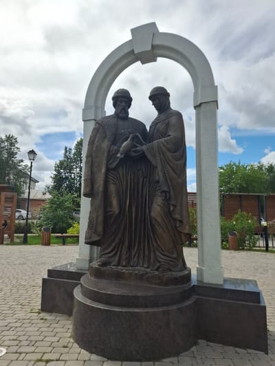 The image shows a bronze statue of two figures standing within a light-colored archway. The figures are dressed in historical clothing. The male figure on the left has a beard and wears a fur-lined coat and a cap. The female figure on the right wears a head covering and a long robe. They are both holding a dove or pigeon. The statue is on a dark granite pedestal. The scene is outdoors, with a paved walkway in the foreground. In the background, there are trees, buildings, a lamppost, benches, and trash cans. The sky is cloudy. This is a monument to St. Peter and St. Paul in Kungur, Russia.