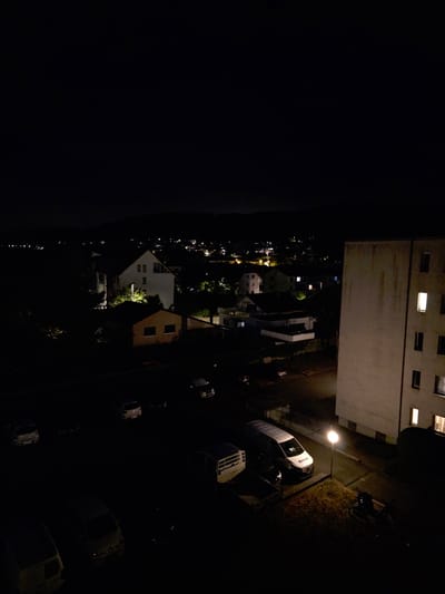 The image shows an aerial view of a residential area at night in Stein, Switzerland. The foreground is dominated by a large, light-colored apartment building on the right, with several windows illuminated. In front of the building, a street light illuminates a patch of grass and pavement. Several vehicles are parked on the street and in a lot, including a white van with "BECKER" written on its side.

In the middle ground, there are several houses and smaller buildings, some with lights on. Trees and foliage are visible, casting shadows in the darkness. The background shows a distant view of more illuminated buildings and houses, suggesting a broader urban or suburban landscape. The sky is dark, with a faint horizon line indicating a distant landscape. The overall scene depicts a quiet night in a residential neighborhood.