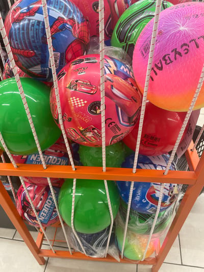 The image shows a close-up, eye-level, vertical shot of a display rack filled with colorful inflatable balls. The rack is made of orange metal bars, and the balls are arranged in rows, separated by white elastic cords. The balls vary in color and design. In the top row, there's a large pink and orange volleyball net ball, a solid red ball, and a green ball. Below that, a green soccer ball with black accents is visible, next to a collection of balls with popular cartoon characters. One ball prominently features Spider-Man, with his red and blue costume and buildings in the background. Another ball is adorned with images of Lightning McQueen from the movie Cars, with the text "LIGHTNING MCQUEEN" clearly visible. Some balls are partially enclosed in clear plastic packaging.
The scene appears to be an indoor retail environment, likely a toy store or a department store section dedicated to sporting goods or children's toys, given the display method and the types of products. There are no people visible, and the lighting suggests an indoor setting. The focus is entirely on the product display.
Visible text includes brand names like "mercan," product-related terms like "VOLLEYBALL," "PVC(WB)," and character names like "SPIDER-MAN" and "LIGHTNING MCQUEEN." There are also product codes and potentially website addresses, such as "spirsport.com" and "www.летсапоулсай.com" which appears to be a Cyrillic transliteration of a Turkish website. The overall impression is one of a vibrant and diverse collection of play balls for children.