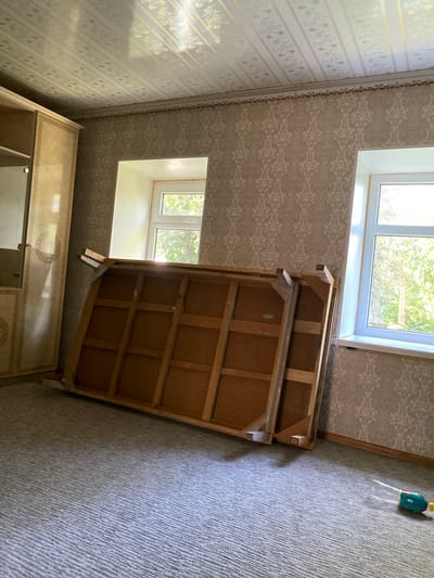 The image depicts an interior view of a room, likely a bedroom or living area, located in Миң-Булак, Kyrgyzstan. The room features patterned wallpaper on the walls and a decorative, possibly paneled, ceiling. Two windows are visible, allowing natural light to illuminate the space. In the foreground, a large, flat wooden object, resembling a disassembled bed frame or a large piece of furniture, is leaning against the wall. A wardrobe or cabinet is positioned to the left of the frame. The floor is covered with a grey, textured carpet. In the lower right corner, a small, colorful toy, possibly a bubble blower, is present on the carpet. The overall impression is that of a room undergoing renovation or being prepared for storage, with furniture partially disassembled and out of place.