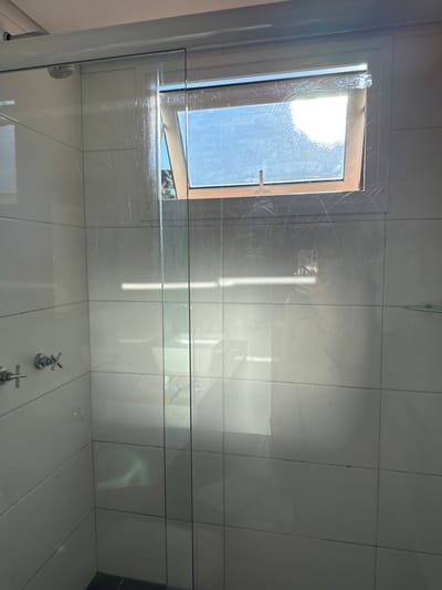 This forensic description details a bathroom shower area located in Canela, Brazil. The scene is dominated by a transparent glass shower enclosure, which features numerous visible water streaks, smudges, and faint scratches across its surface. To the left, within the shower, the walls are covered in rectangular white tiles, upon which a chrome-finished shower head is mounted in the upper left corner, and two chrome cross-shaped faucet handles are visible on the lower left.
Above the shower enclosure, a top-hinged window with a light-colored frame is partially open, revealing a clear blue sky and distant green foliage, indicating an outdoor view. Bright sunlight streams through this window, creating strong reflections on the glass. Through the glass, on the lower right side, a reflection of a white bathroom sink or vanity with a faucet is discernible, positioned behind the camera's viewpoint. A clear reflection of a person holding a smartphone, the likely photographer, is also evident on the right side of the glass enclosure. The floor within the shower appears to be a dark color. No other individuals or direct interactions are visible in the scene.