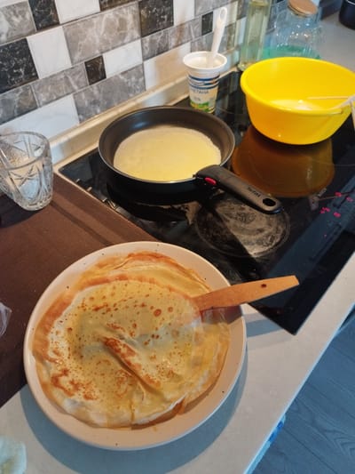 In the city of Lymanka, Ukraine, an indoor scene depicts the process of making crepes. A stovetop, possibly electric, is visible, with a frying pan containing a thin batter. To the right, a bright yellow bowl holds more batter, and a white cup with a lid and spoon is nearby, possibly containing sour cream. A glass of water sits to the left, and a stack of finished crepes on a plate is in the foreground. A wooden spatula rests on the plate, touching one of the crepes. The background features a tiled backsplash and kitchen counter. The lighting suggests a domestic setting, possibly during daylight hours.