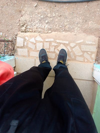 In El Maco, Venezuela, a first-person perspective shows a person wearing black pants and dark sneakers with yellow accents, standing on a mosaic tiled path. The path is bordered by dirt and rocks, with a glimpse of a red object and a metal railing to the left. To the right, a section of a green wall is visible.