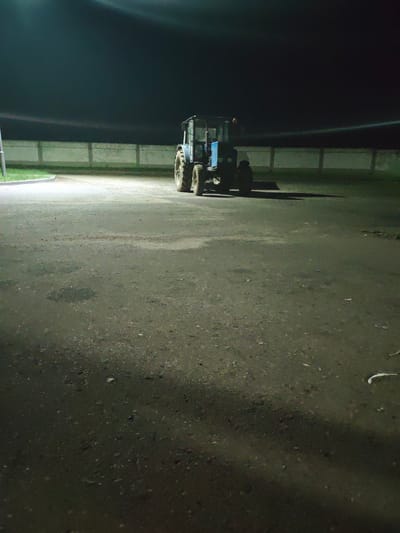 The image depicts an outdoor scene at night, illuminated by artificial lights, in the vicinity of Скідзельскі сельскі Савет, Belarus. The ground is an asphalt or gravel surface, appearing somewhat worn and wet in places, with some debris scattered across it. In the mid-ground, a blue tractor is parked facing towards the right of the frame. The tractor has large rear wheels and a cabin structure. Behind the tractor, a light-colored concrete fence runs horizontally across the image, topped with what appears to be barbed wire. The background is dark, indicating the absence of natural light. The overall impression is of an industrial or agricultural area at rest during nighttime.