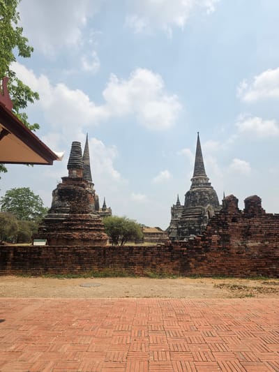 The image shows the ruins of Wat Yai Chaimongkol in Phra Nakhon Si Ayutthaya City Municipality, Thailand. The photo is taken at ground level with the camera rotated to place the main structures vertically. Several brick chedis are visible, showing signs of age and decay. A brick pathway, also worn, leads away from the structures. A partly visible roof of a modern structure and some foliage are in the foreground. The sky is visible in the background with scattered clouds.