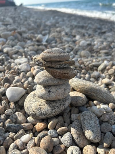 A close-up, eye-level, vertical shot features a stack of five smooth, grey and brown stones balanced on a pebble-strewn beach. The stones are weathered and have distinct lines and textures. The background is softly focused, showing the continuation of the pebble beach stretching towards the ocean, where gentle waves are breaking under a bright, hazy sky. The scene is devoid of any people or specific landmarks of Tolbazikha, Russia. The lighting suggests it is daytime.