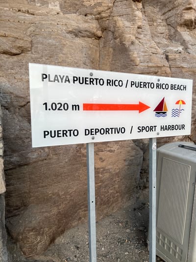 The image shows a directional sign in Mogán, Spain, indicating that the Playa Puerto Rico beach and the Puerto Deportivo sport harbor are 1.020 meters in the direction indicated by the red arrow. The sign is attached to a metal frame. The background shows a rocky terrain.