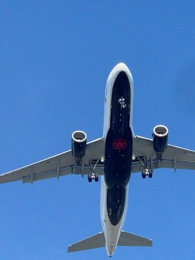 An airplane is captured from below, ascending against a clear blue sky. The underside of the fuselage is predominantly black, featuring a red circular logo with a maple leaf emblem, identifying it as Air Canada. Two jet engines are visible, one on each wing, with a white ring around the intake of the visible engine. The landing gear is deployed, with wheels down, suggesting the aircraft is either taking off or landing. The scene is consistent with an airplane in flight over Richmond, Canada, given Air Canada's presence at Vancouver International Airport (YVR), a major airport serving the city.