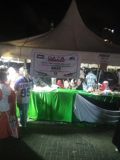The image captures a nighttime outdoor event within the Makadara/Mji Wa Kale ward, Kenya, under a large white peaked tent. A prominent banner is centrally displayed, featuring the text "GAZA MEDICAL APPEAL," a Palestinian flag, and the "Yadul Ulya" logo, with the message, "Yadul-Ulya humbly invites you to assist in raising Medical Funds for GAZA." To the right of this banner, a smaller sign indicates "LIPA NA MPESA 150770 TreadGaza." Below the banner, two tables are positioned, covered primarily with green fabric. The table on the right also displays white and black fabric, completing the colors of the Palestinian flag. Various items, including food trays and bottled water, are arranged on these tables. Several individuals are present under the tent's artificial lighting. In the left foreground, a person is visible wearing an orange and white garment. Slightly behind them, another individual is seen from the back, wearing a blue and white shirt with "MENGO 95" printed on it. Further right, behind the table, a person wearing a red head covering is partially visible. The ground is paved with interlocking blocks. In the background, indistinct urban buildings and a structure resembling a minaret with a green top are faintly discernible against the dark sky.