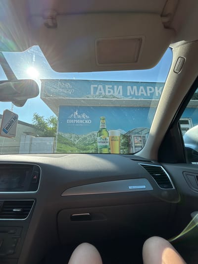 An eye-level shot, likely taken by a passenger, looks out from the interior of a car through a sun-drenched windshield. The image is oriented vertically (rotated 90 degrees clockwise from typical viewing).
Inside the car, the beige sun visor is visible on the upper left, bearing an "AIRBAG" label. Part of the car's dashboard, air vents, and central console are seen on the right, featuring a "Quattro" badge and a digital display showing "1:20". Below, several buttons are visible including "SCAN", "TEL", "CAR", and "SETUP", along with a hazard light button. A "mobile.bg" air freshener hangs from the rearview mirror in the lower center. A bare human leg is partially visible on the far right. The windshield has some smudges and significant lens flare from the bright sun.
Outside the car, the primary subject is a large billboard advertising "ПИРИНСКО" (Pirinско) beer, a Bulgarian brand, with text "светло" (svetlo - light). The billboard also displays "ГАБИ МАРК" (Gabi Mark) and features an image of mountains and beer bottles/glasses. The background reveals a clear blue sky, some green trees, and parts of buildings. The bright, clear weather suggests it's daytime in Rakovski, Bulgaria, as indicated by the context and Bulgarian text.